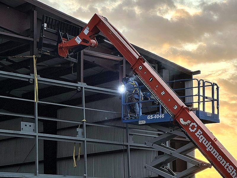Welder on a scissor lift beside a telehandler at sunrise