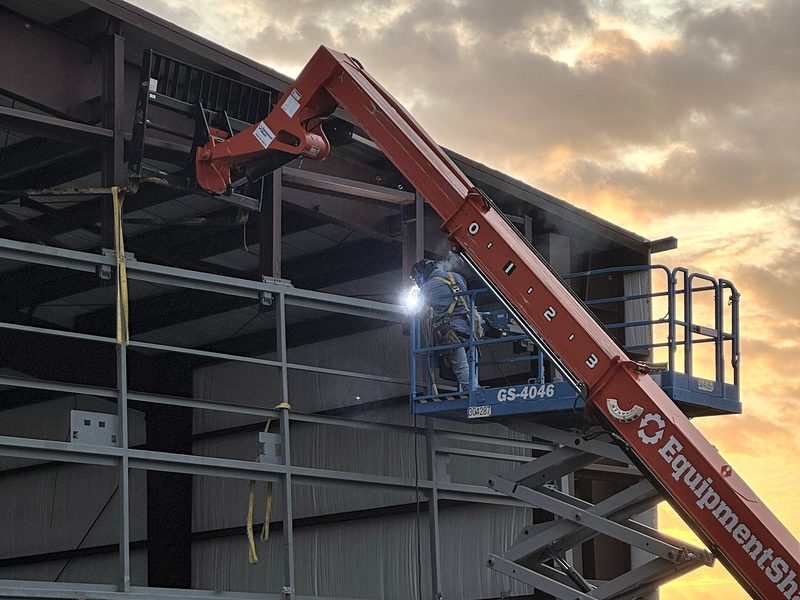 Welder on a scissor lift beside a telehandler at sunrise